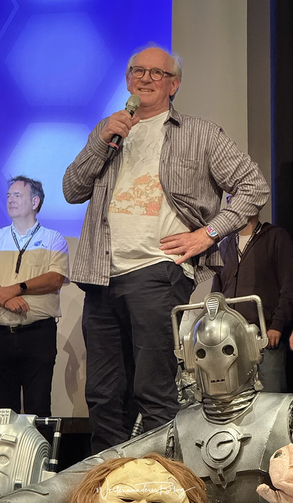 Peter Davison mit Mikrofon auf der Bühne der TimeLash X, davor eine Cyberman-Figur